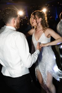 A man and woman in formal attire dance closely at a lively indoor event, illuminated by dramatic lighting with people in the background—a moment beautifully captured through documentary photography.