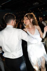 A man in a white suit and a woman in a sparkly white dress dance together at a lively indoor event, captured in an editorial photography style, with other people visible in the background.