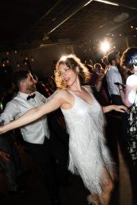 A woman in a white fringed dress dances energetically at a lively indoor party in Toronto, surrounded by other people. Bright lights shine in the background, adding to the festive atmosphere—capturing a vibrant moment of documentary photography.