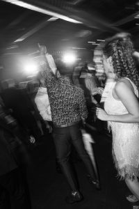 Black and white photo of people dancing at a party, captured with a keen documentary eye. A person in a patterned shirt raises an arm, while another in a fringed dress faces away. Bright lights and motion blur add energy to the lively scene, emphasizing the photographer’s editorial style.