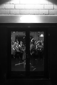 A group of people are seen through glass doors at night, gathered closely together, some raising their arms or holding objects, illuminated by indoor lighting. Captured in black and white documentary photography, the scene has a candid quality that draws the viewer into the moment.