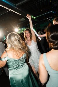 A woman in a sparkly white dress dances energetically with her arm raised at a lively indoor party, surrounded by others in green dresses under festive lighting, as a photographer captures the vibrant atmosphere through their lens—a true celebration of Toronto’s dynamic party scene.