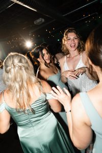 Four women in light green and white dresses are smiling and dancing together at a party in Toronto, captured in bright lighting against a dark, festive background that gives the scene an editorial and documentary feel.