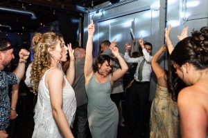A group of people dressed in semi-formal attire dance energetically at an indoor party in Toronto, raising their arms and smiling under colorful lights, capturing a lively moment fit for editorial or documentary coverage.