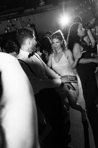People dressed formally are dancing at a party, reminiscent of a lively wedding celebration. A woman in a sparkly dress is looking at a man in a white shirt and bow tie, while the scene is lit by a bright light in the background, capturing a perfect moment for any documentary photographer.
