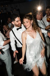 A man in a tuxedo and a woman in a sparkly dress dance and smile together at a lively indoor party, captured in beautiful documentary photography, with other joyful guests in the background.