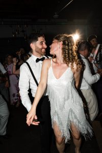 A couple dressed in formal attire smiles and dances closely at a lively party in Toronto, captured by an editorial photographer, surrounded by other people enjoying the celebration.