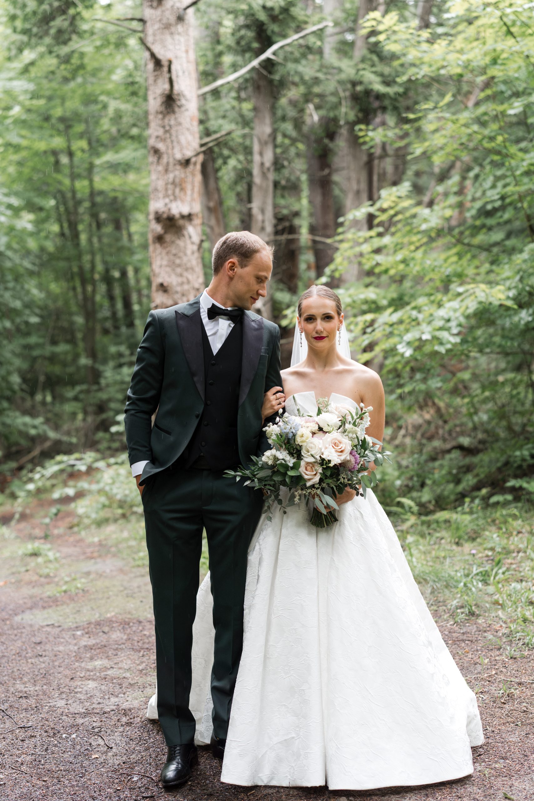 A bride in a white gown holding a bouquet stands arm-in-arm with a groom in a dark suit and bow tie on a forested path, surrounded by green trees, capturing an editorial photography moment onto the tranquil woodland setting.