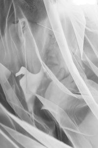 A black and white photo of a couple close together, partially obscured by a sheer veil, sharing an intimate, tender moment—capturing the timeless romance of a wedding in an editorial style.