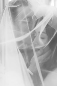 A black-and-white photo, captured by a wedding photographer, shows a couple under a sheer veil. One person gently holds the other’s face and kisses their forehead; both have their eyes closed, sharing an intimate, tender moment.