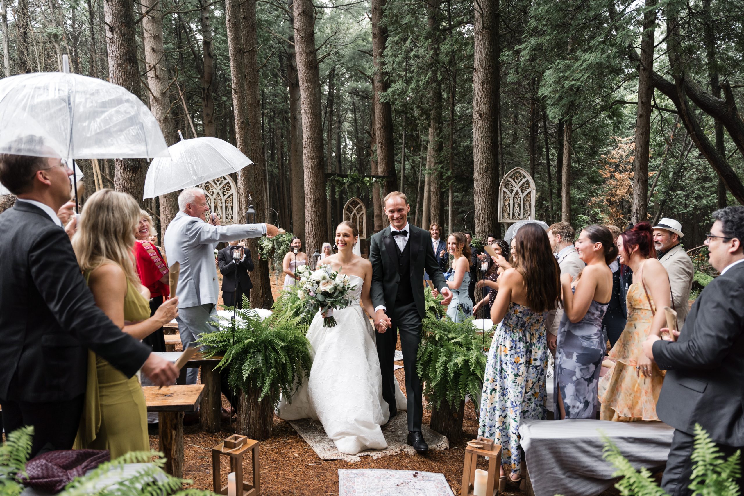 A bride and groom walk down the aisle in a forest wedding, smiling as guests stand, cheer, and hold umbrellas in the rain. The scene is surrounded by tall trees and lush green ferns, capturing the magic of wedding photography as sunlight filters onto the happy couple.