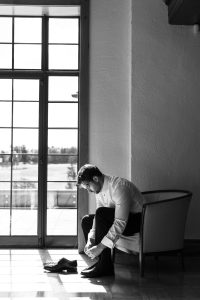 A man sits on a chair by a large window, putting on dress shoes as sunlight streams in—an elegant black and white moment often captured by wedding photographers Toronto cherishes.