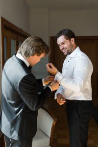 Two men in formal attire prepare for an event. One, in a white dress shirt, smiles as the other, in a black suit, helps adjust his cufflink—captured beautifully by Wedding Photographers Toronto.
