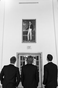 Three men in suits stand with their backs to the camera, looking up at a large portrait above an EXIT sign; a classic black and white image reminiscent of Wedding Photographers Toronto style.