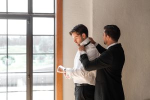 Two men in suits stand by a window as sunlight streams in. One adjusts the other’s bow tie—a perfect moment for Wedding Photographers Toronto to capture the warm, soft atmosphere.