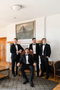 Four men in black tuxedos pose in a living room; three stand while one sits on a wooden armchair. Captured by Wedding Photographers Toronto, the formal scene features a mountain painting above the fireplace.