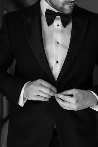 A man in a tuxedo fastens his jacket button. He wears a white dress shirt with black buttons and a bow tie. Captured by Wedding Photographers Toronto, the black-and-white image shows his face partially out of view.