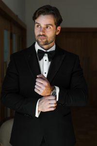A man in a black tuxedo adjusts his cufflink while looking to the side. He has short hair, a beard, and stands in a dimly lit room—perfect inspiration for Wedding Photographers Toronto.