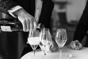 A person in a suit pours sparkling wine into champagne glasses on a table, with another person nearby—a timeless black and white moment captured by Wedding Photographers Toronto.