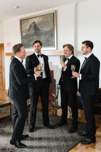 Four men in black tuxedos stand together indoors, smiling and holding glasses of white wine. Wedding Photographers Toronto could capture this elegant celebration in a well-lit room with a painting and fireplace behind them.
