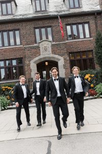 Four men in black tuxedos are joyfully walking outside a brick building with large windows and flowers, smiling and laughing together—perfectly captured by wedding photographers Toronto.