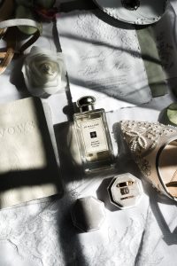 Flat lay of wedding items on lace fabric, beautifully styled by Wedding Photographers Toronto, featuring a Jo Malone perfume bottle, vow book, pearl shoe, white rose, rings, and invitations softly lit by sunlight.
