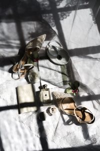 Flat lay of bridal items—lace heels, vow book, perfume, hexagonal ring box, white flower, earrings, and a small mirror—styled on lace fabric in sunlight. A dreamy setup captured by Wedding Photographers Toronto.