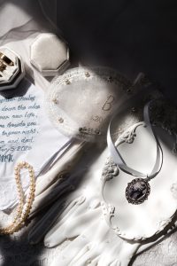 Vintage bridal accessories—white gloves, a pearl bracelet, embroidered fabric, and a cameo necklace—are beautifully arranged for Wedding Photographers Toronto on a reflective tray in soft sunlight.