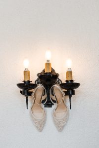 A pair of elegant white bridal shoes hangs from a black, three-armed wall sconce with lit candle-style bulbs—a perfect detail for Wedding Photographers Toronto to capture against the white textured wall.