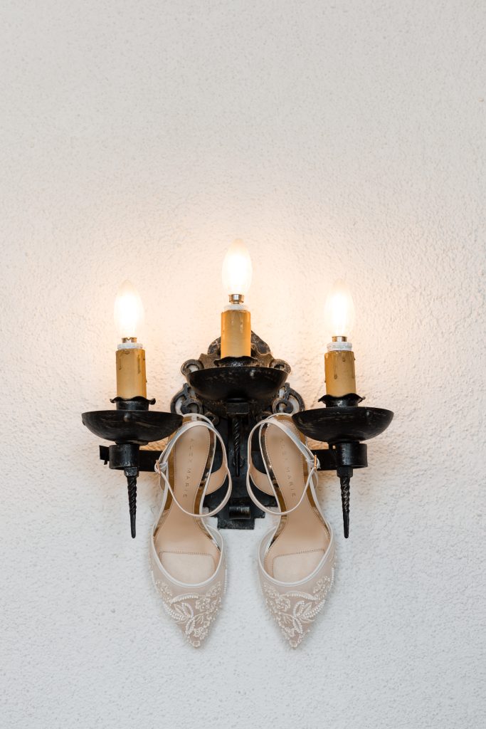 A pair of elegant white bridal shoes hangs from a black, three-armed wall sconce with lit candle-style bulbs—a perfect detail for Wedding Photographers Toronto to capture against the white textured wall.