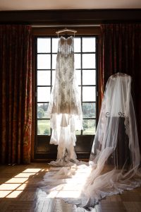 A white lace wedding dress hangs in front of a sunlit window, with a long, sheer veil draped nearby—perfect inspiration for Wedding Photographers Toronto seeking warm light and elegant detail.