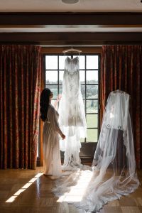 A woman in a robe stands by a window, touching a white lace wedding dress glowing in sunlight. Wedding Photographers Toronto capture the moment, as rich red curtains frame the veil-draped dress form nearby.