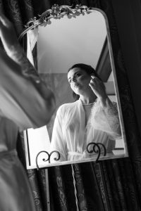A woman in a white robe adjusts an earring while looking into an ornate mirror, her reflection clear. Captured by Wedding Photographers Toronto, the black-and-white scene features patterned curtains in the background.