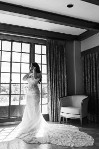 A bride in a lace gown stands by large windows, adjusting her earrings, with sunlight streaming in. An empty chair sits nearby; captured in black and white by Wedding Photographers Toronto.