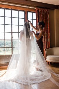 A woman in a brown dress adjusts the veil of a bride standing by a sunlit window, as Wedding Photographers Toronto capture the moment, highlighting her white gown and lace veil.