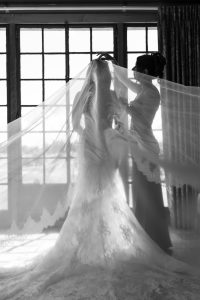 A bride stands in front of large windows as another woman arranges her veil. Light streams through the lace, creating a soft, elegant scene in black and white—perfectly captured by Wedding Photographers Toronto.