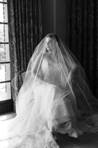 A bride in a lace wedding dress and veil sits by a window, looking thoughtfully outside. Sunlight softly illuminates her face through the sheer veil—captured artfully by Wedding Photographers Toronto.