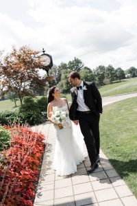 A bride and groom walk hand in hand on a garden path, smiling at each other. Captured by Wedding Photographers Toronto, the bride holds a bouquet as a clock stands in the background among trees and flowers.