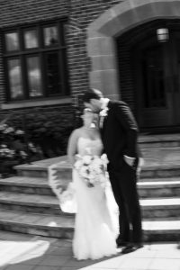 A black and white, blurry photo by Wedding Photographers Toronto shows a couple in wedding attire on stone steps outside a brick building as the groom leans down to kiss the bride holding a large bouquet.