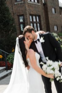 A bride and groom share a joyful kiss outside, captured perfectly by Wedding Photographers Toronto. The bride holds a bouquet of white flowers, wearing a veil, while the groom dons a black suit with a boutonniere.