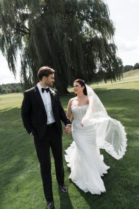 A bride in a white lace gown and a groom in a black tuxedo walk hand in hand on green grass with a large willow tree in the background, smiling at each other—captured perfectly by Wedding Photographers Toronto.