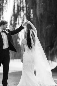 A groom twirls his bride, who wears a long veil and strapless gown, outdoors under tall trees. Wedding Photographers Toronto capture the timeless moment in stunning black and white.