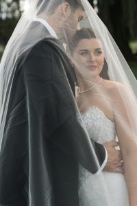 A bride and groom stand close together under a sheer veil outdoors. Captured by Wedding Photographers Toronto, the groom wears a black suit, and the bride smiles softly in her white lace dress and pearl necklace.