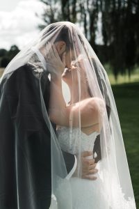 A bride and groom kiss under a veil outdoors, with sunlight illuminating their faces and soft greenery in the background—captured beautifully by top Wedding Photographers Toronto.