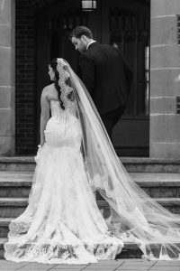 A bride in a long lace wedding dress and veil stands beside a groom in a suit as they ascend stone steps outside a building, captured from behind in black and white by Wedding Photographers Toronto.