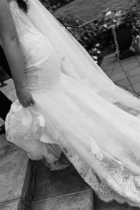 Close-up of a bride in a lace wedding dress walking up stone steps, holding her train. Captured by Wedding Photographers Toronto, the black and white image features flowers and greenery in the background.