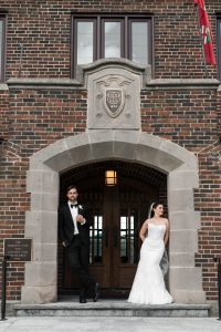 A bride and groom pose outside a brick building under an archway, the groom leaning on one side and the bride on the other, both looking at each other and smiling—captured beautifully by wedding photographers Toronto.