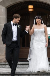 A bride in a white lace dress and a groom in a black tuxedo hold hands while walking down stone steps—captured beautifully by top Wedding Photographers Toronto—outside a building with an arched doorway.