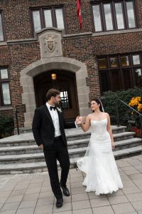 A bride in a white gown and groom in a black tuxedo hold hands, smiling at each other, as Wedding Photographers Toronto capture their walk outside a brick building with stone steps and an arched doorway.