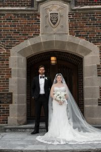 A bride in a white dress and veil stands beside her groom in a black tuxedo, posing outside a brick building with an arched doorway—a classic scene captured by top Wedding Photographers Toronto.